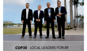 Quatro homens vestidos de terno posam em um palco ao ar livre com a inscrição “COP30 Local Leaders Forum”, tendo ao fundo um cenário arborizado e ensolarado.