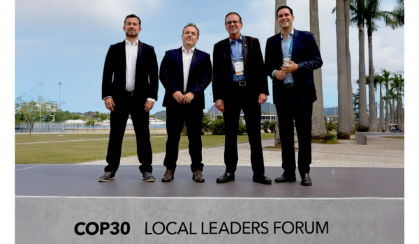 Quatro homens vestidos de terno posam em um palco ao ar livre com a inscrição “COP30 Local Leaders Forum”, tendo ao fundo um cenário arborizado e ensolarado.