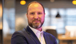 Homem de barba e cabelo curto posa com os braços cruzados, usando terno azul e camisa branca, em um ambiente de escritório moderno e iluminado.