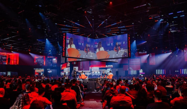 Grande evento corporativo em um auditório moderno, com palco central iluminado em tons de vermelho e azul. Há várias pessoas assistindo a uma conversa entre palestrantes sentados em poltronas laranja, transmitida em um telão suspenso acima do palco.