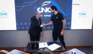 Dois homens apertam as mãos em frente a uma tela com o logotipo da CNC, durante uma reunião formal. Um deles veste terno e gravata, e o outro, roupa casual.