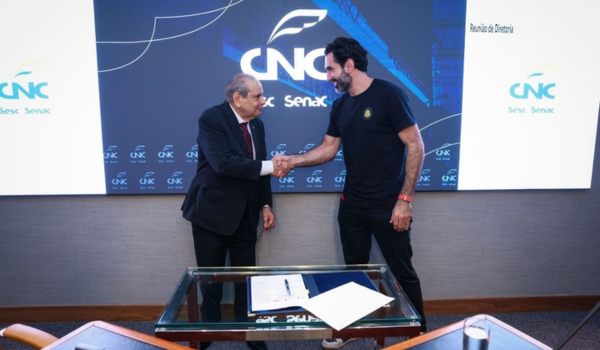 Dois homens apertam as mãos em frente a uma tela com o logotipo da CNC, durante uma reunião formal. Um deles veste terno e gravata, e o outro, roupa casual.