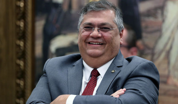 Homem de terno cinza e gravata vermelha sorri com os braços cruzados, em um ambiente formal com quadro decorativo ao fundo.