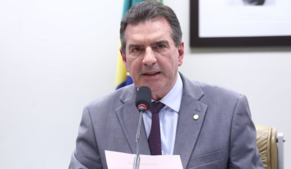 Homem de terno cinza e gravata roxa fala ao microfone durante uma sessão oficial, segurando um papel nas mãos. Ao fundo, há uma bandeira do Brasil parcialmente visível e um quadro na parede.