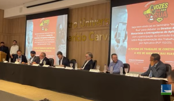 Painel de debate com vários participantes sentados à mesa em auditório, durante o seminário “Vozes da Rua”, que discute os direitos de motoristas e entregadores de aplicativo.