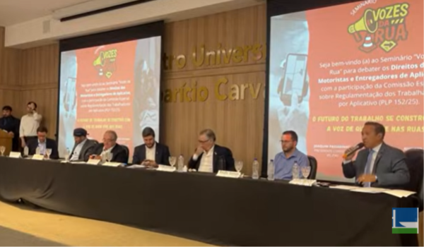 Painel de debate com vários participantes sentados à mesa em auditório, durante o seminário “Vozes da Rua”, que discute os direitos de motoristas e entregadores de aplicativo.