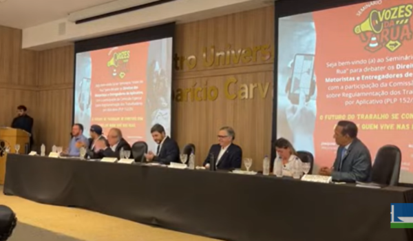 Painel de debate com vários participantes sentados à mesa em auditório, durante o seminário “Vozes da Rua”, que discute os direitos de motoristas e entregadores de aplicativo.