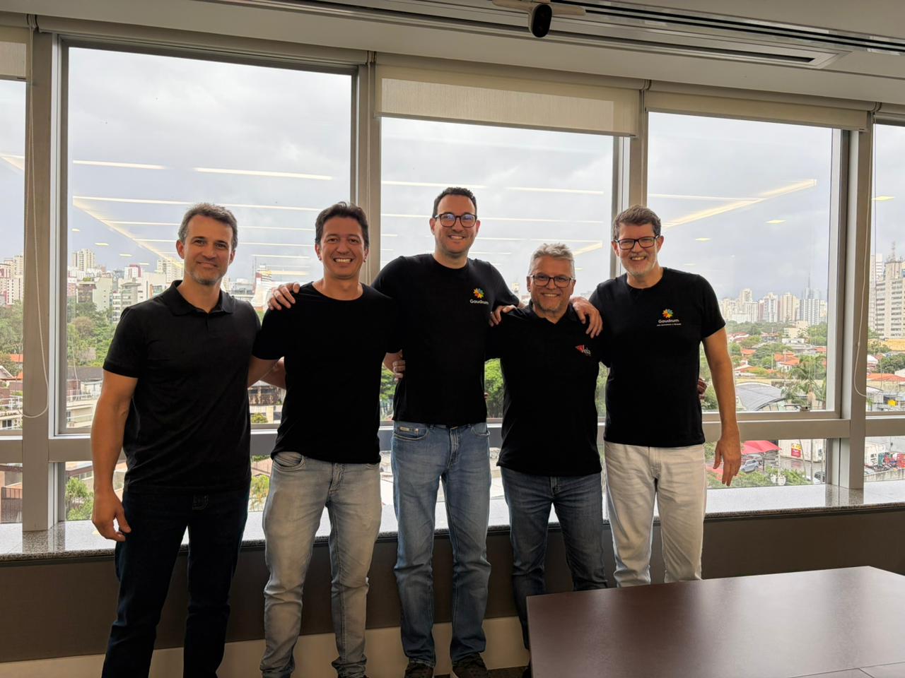 Cinco homens em pé, lado a lado, sorrindo em frente a uma janela ampla com vista urbana ao fundo.
