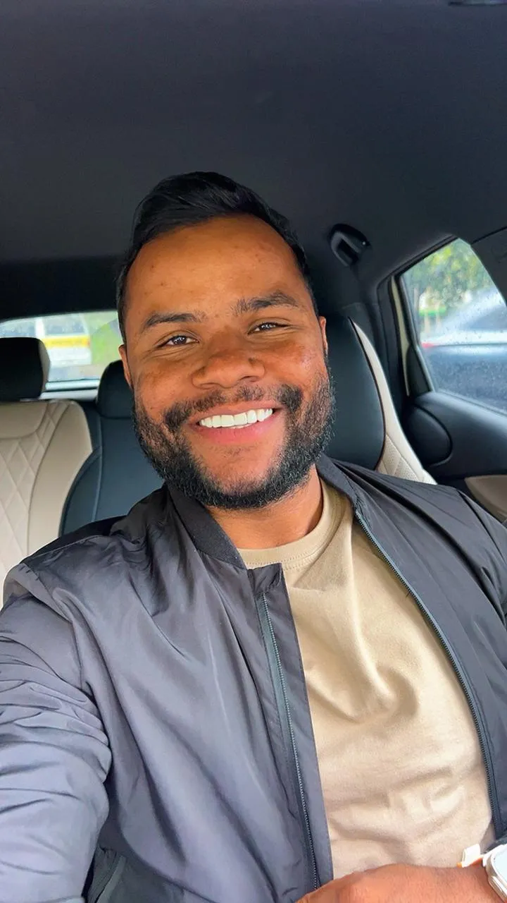 Homem sorrindo dentro de um carro, usando jaqueta escura e camiseta bege.