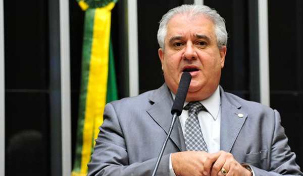 Homem de cabelo grisalho, em terno cinza e gravata, fala ao microfone em um plenário; ao fundo há uma faixa verde e amarela.