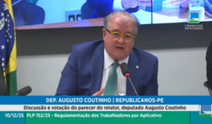 Homem de terno azul e óculos fala ao microfone em audiência na Câmara dos Deputados, com banner na tela sobre discussão e votação de parecer.