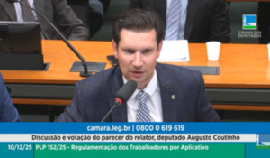 Deputado fala ao microfone durante audiência na Câmara dos Deputados, com banner na tela sobre votação e regulamentação de trabalhadores por aplicativo.