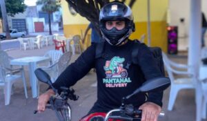 Entregador de moto usando capacete preto e camiseta com o logo 'Pantanal Delivery', sentado em uma motocicleta em uma área externa com mesas e cadeiras ao fundo."