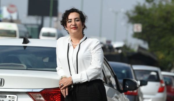 Mulher de cabelos curtos e cacheados, vestindo camisa branca e calça escura, posa apoiada na traseira de um carro prata em uma rua movimentada com vários veículos ao fundo.