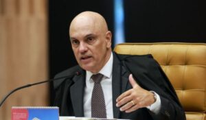Ministro do Supremo Tribunal Federal fala durante sessão, sentado em uma cadeira de couro, usando terno e toga, com a mão levantada e um microfone à frente.