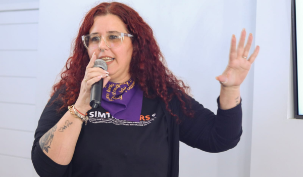 Mulher de cabelos cacheados e avermelhados fala ao microfone enquanto gesticula com a mão levantada, usando óculos e uma camiseta preta.