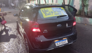 Carro preto visto por trás em rua molhada, com adesivos de propaganda no vidro traseiro e placa brasileira.
