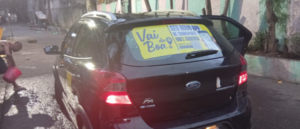 Carro preto visto por trás em rua molhada, com adesivos de propaganda no vidro traseiro e placa brasileira.