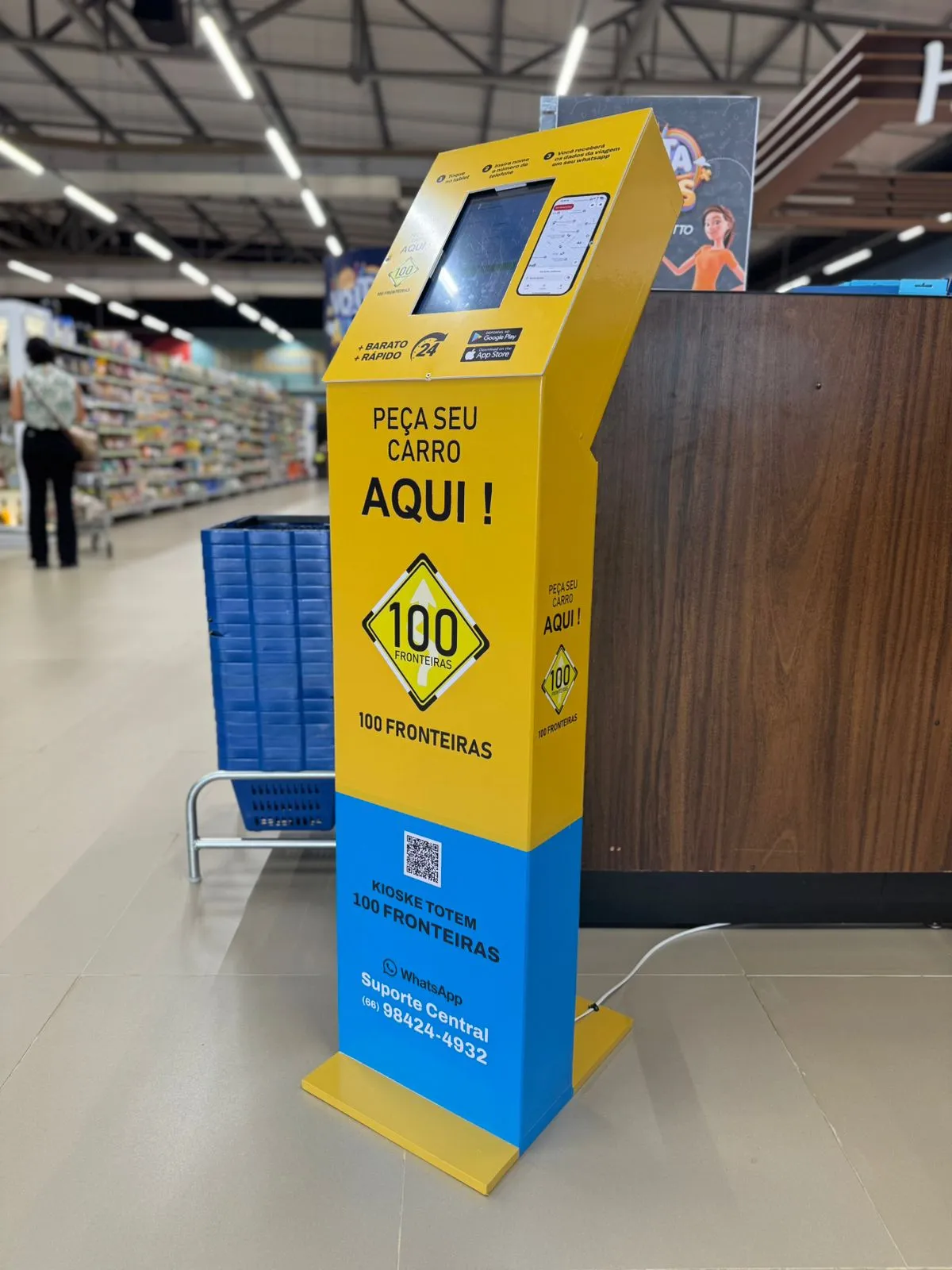 Totem amarelo com a mensagem “Peça seu carro aqui” e logotipo 100 Fronteiras em área interna de supermercado, ao lado de carrinhos de compras.