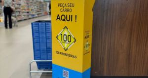 Totem amarelo com a mensagem “Peça seu carro aqui” e logotipo 100 Fronteiras em área interna de supermercado, ao lado de carrinhos de compras.