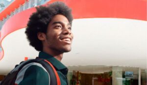 Jovem sorridente com mochila nas costas, em frente a um prédio moderno com detalhes em vermelho.