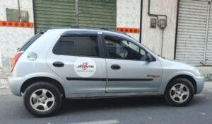Carro prata modelo Chevrolet Celta estacionado na rua, com um adesivo circular de aplicativo de transporte na porta traseira e a inscrição "OFFROAD" na porta dianteira.