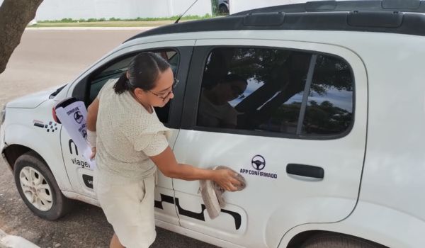 Mulher adesivando a porta de um carro branco com a logomarca 'App Confirmado'. Ela segura papéis sob o braço enquanto aplica o adesivo na lateral do veículo estacionado sob a sombra de uma árvore