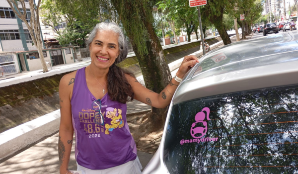 Mulher sorridente de cabelos grisalhos presos, usando regata roxa esportiva, em pé ao lado de um carro prata estacionado em uma rua arborizada; no vidro traseiro do carro há um adesivo rosa com o texto “@mamydriver”.
