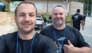 Dois homens sorrindo em uma selfie ao ar livre, usando camisetas pretas e crachás azuis; o homem à direita faz sinal de positivo com o polegar, enquanto pessoas e veículos aparecem ao fundo em um ambiente de evento.