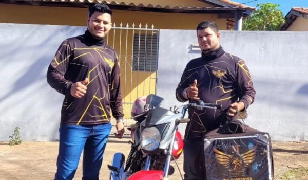 Dois homens sorridentes vestindo uniformes pretos com detalhes dourados posam ao lado de uma motocicleta vermelha. O homem à direita segura uma mochila de entregas personalizada com o mesmo logotipo da farda.