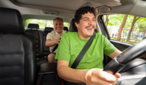 Motorista sorridente dirigindo carro com passageiro no banco de trás, ambos usando cinto de segurança.