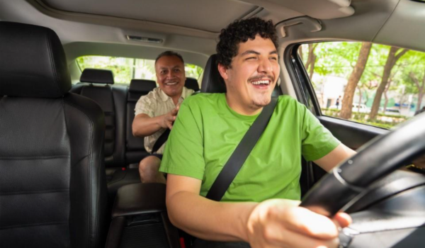 Motorista sorridente dirigindo carro com passageiro no banco de trás, ambos usando cinto de segurança.