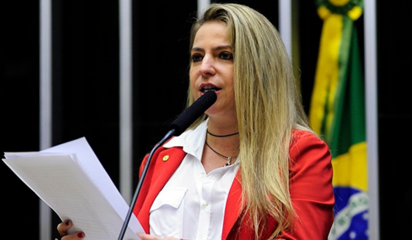 Mulher de blazer vermelho discursa na tribuna da Câmara dos Deputados, segurando folhas de papel.