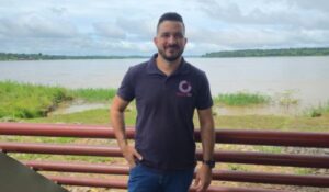 Homem em pé apoiado em corrimão vermelho à beira de um rio largo, usando camiseta polo com logotipo Connect Trip, com vegetação ao fundo e céu nublado