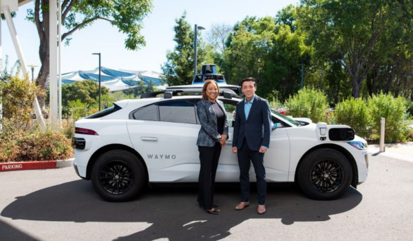 Duas pessoas em pé ao lado de um carro autônomo branco da Waymo estacionado em área externa arborizada