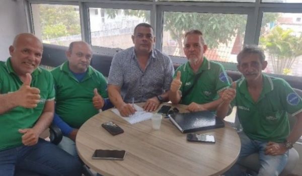Cinco homens sentados ao redor de uma mesa assinando documentos, alguns fazendo gesto de positivo, em ambiente interno com janelas ao fundo.