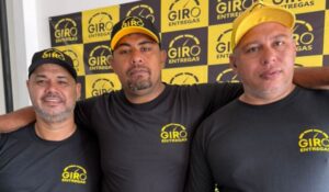 Três homens com camisetas e bonés pretos com o logotipo Giro Entregas posam juntos diante de um painel da marca