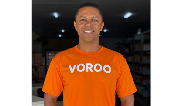Homem sorrindo vestindo camiseta laranja com a palavra “VOROO” em ambiente interno com prateleiras ao fundo