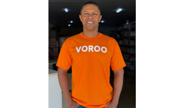 Homem sorrindo vestindo camiseta laranja com a palavra “VOROO”, em ambiente interno de loja