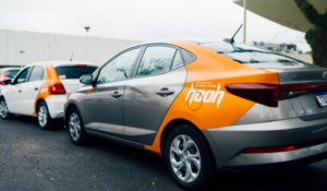 Carros com adesivos do aplicativo HooH estacionados em fila em área urbana, com destaque para veículo cinza e laranja.