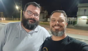 Dois homens sorrindo tiram uma selfie à noite em uma rua iluminada, com casas ao fundo.