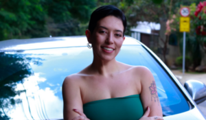 Mulher sorrindo com braços cruzados em frente a um carro estacionado em rua arborizada, usando top verde e com tatuagens visíveis nos braços
