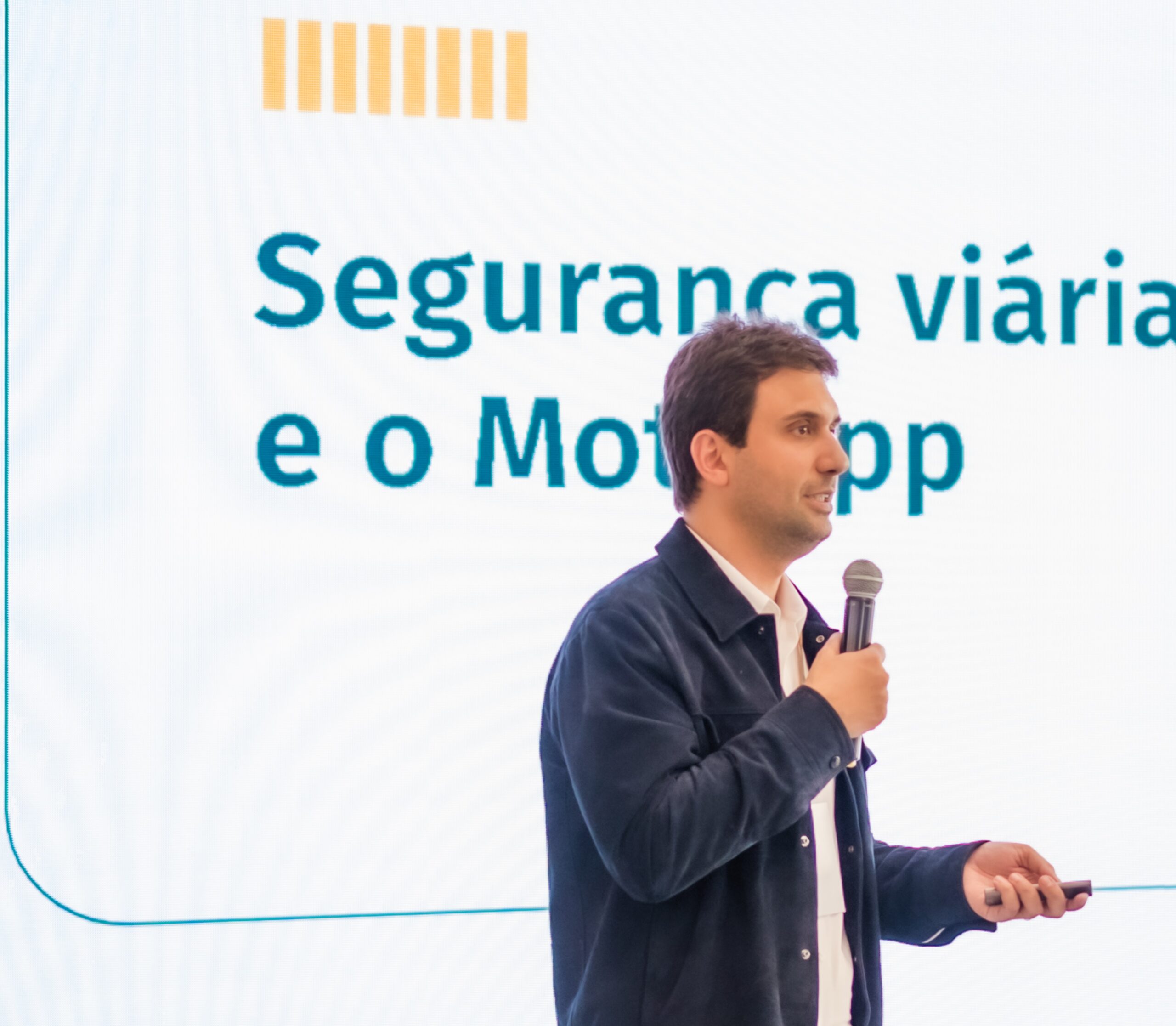Homem segurando microfone enquanto faz apresentação diante de slide com o texto “Segurança viária e o Mot…” ao fundo