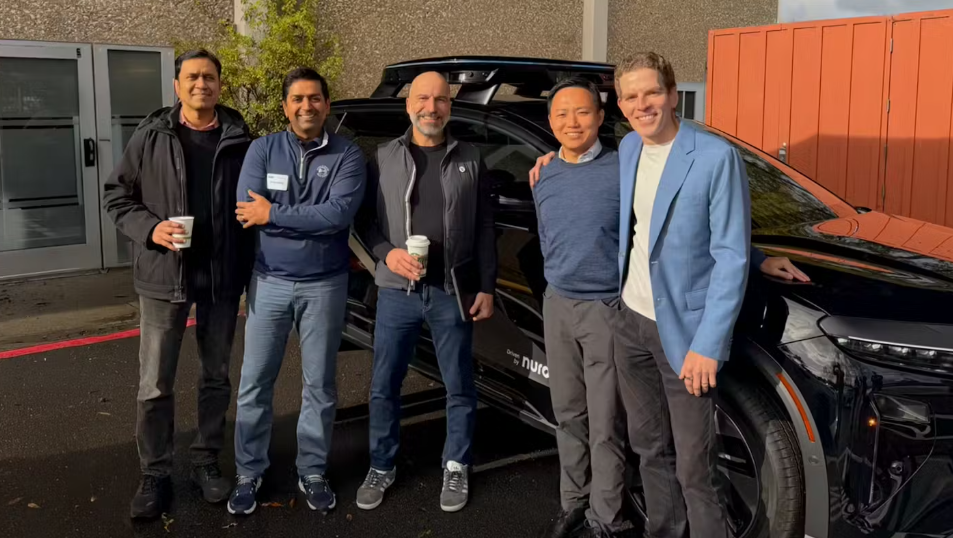 Cinco homens posam sorrindo ao lado de um veículo preto da Nuro estacionado ao ar livre.