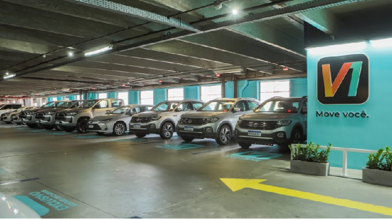 Fila de carros estacionados em garagem coberta com vagas sinalizadas e parede com logotipo V1 e frase “Move você.”