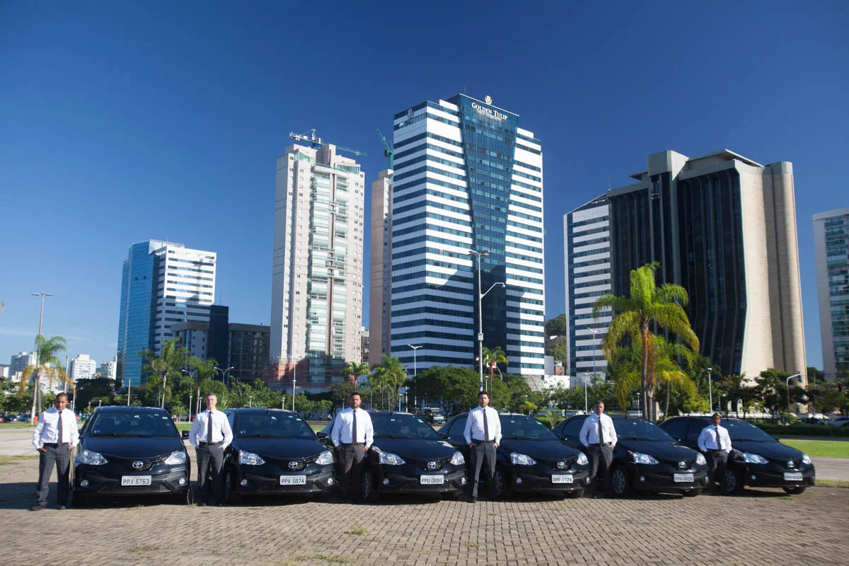 Motoristas uniformizados ao lado de carros pretos alinhados em frente a prédios comerciais modernos