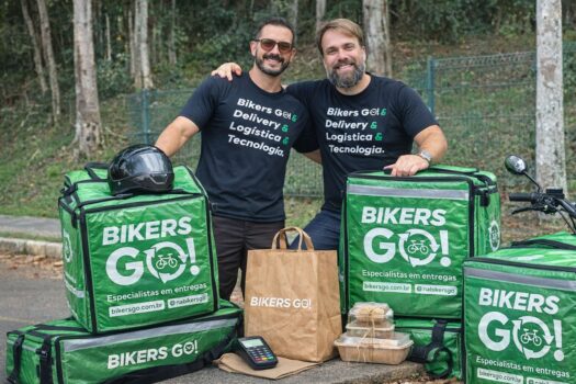 Dois homens sorrindo ao lado de mochilas térmicas verdes de entrega com logotipo Bikers Go, com sacolas de papel e embalagens de comida em primeiro plano.