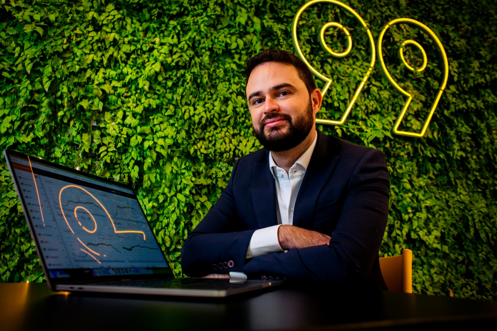 Homem de terno sentado à mesa com laptop exibindo gráficos, diante de parede verde com iluminação neon em formato de interrogação