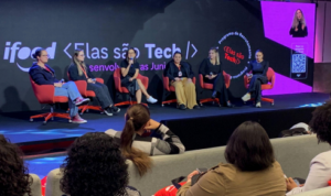 Painel com mulheres debatendo tecnologia em palco do evento “Elas são Tech”, com plateia ao fundo e identidade visual do iFood.