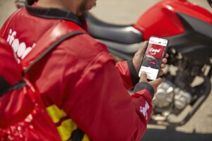 Entregador do iFood com mochila térmica segurando celular com aplicativo aberto ao lado de motocicleta.